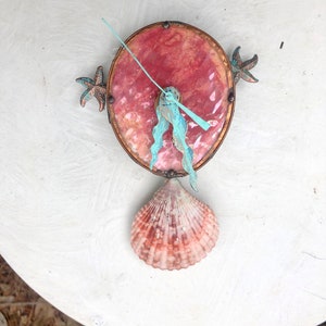 May include: A handcrafted wall clock with a seashell theme. The clock features a round, reddish-pink face with bronze-colored trim. Two bronze starfish adorn the sides, and a seashell hangs below. The clock hands are light blue.