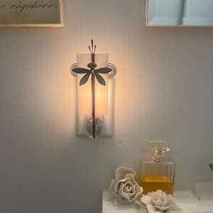 May include: A plug-in wall night light featuring a rectangular frosted glass panel illuminated from behind. A dark metal dragonfly ornament is centered on the glass, flanked by two copper-colored wire spirals. A clear glass bead sits at the base.