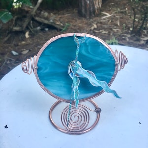 May include: A teal blue stained glass clock with a copper wire frame and stand. The clock face features a crescent moon and a stylized wave design.