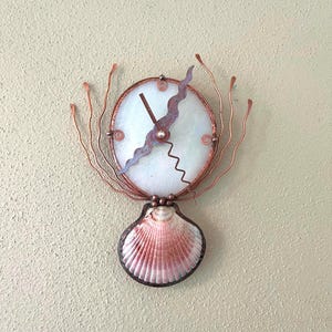 May include: A unique wall clock featuring a white face with copper wire accents. The clock has a seashell base with pink and white hues. The clock's design includes a copper wire frame and decorative elements, creating a coastal theme.
