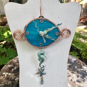May include: A turquoise blue stained glass clock with copper wire accents and a seahorse and starfish charm hanging from the bottom. The clock is displayed on a white wooden stand.