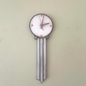 May include: A wall clock featuring a circular white glass face with copper-toned hands, including one wavy hand. The clock has a long, vertical, rectangular white glass body extending downward, accented with copper metal trim and small circular details.