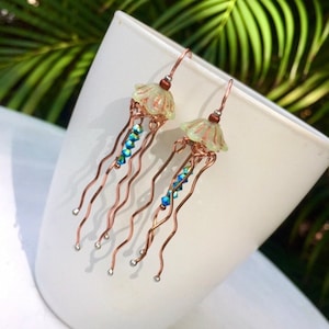 May include: A pair of dangle earrings featuring a light green glass jellyfish charm with copper wire tentacles and blue iridescent beads.