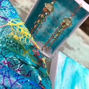 May include: A pair of long, dangling earrings with green jellyfish charms and copper chain links. The earrings are displayed in a small, white box with a blue interior.