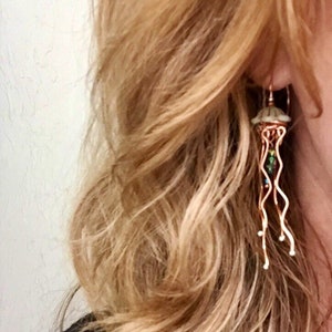 May include: Copper wire jellyfish earrings with a white and green glass bead body. The earrings are dangling from the earlobe.
