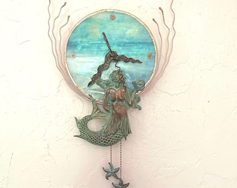 May include: A circular wall clock featuring a teal and blue ocean-inspired face with copper-colored wire accents extending outward. A bronze-toned mermaid figure is attached to the bottom, holding two dangling starfish charms on small chains.