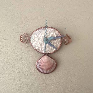 May include: A handcrafted wall clock featuring a round, textured face with copper wire accents. The clock has blue hands and a decorative seashell at the bottom. The clock's design includes copper wire spirals on either side.