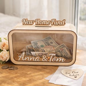Può includere: Un salvadanaio in legno con un fronte trasparente, pieno di contanti. La scatola è etichettata "New Home Fund" e "Anna & Tom". Un piccolo cartello a forma di cuore recita "For Our Future".