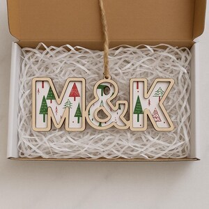 May include: A wooden hanging ornament featuring the letters M and K connected by an ampersand. The letters have a light wood frame and a white interior surface decorated with a repeating pattern of small red and green Christmas trees.