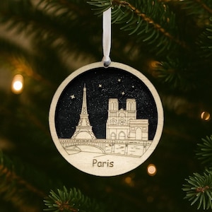 May include: Circular wooden Christmas ornament with a laser-cut design of the Eiffel Tower and Notre Dame Cathedral in Paris, France, against a starry night sky. The word "Paris" is at the bottom. It hangs from a white ribbon.