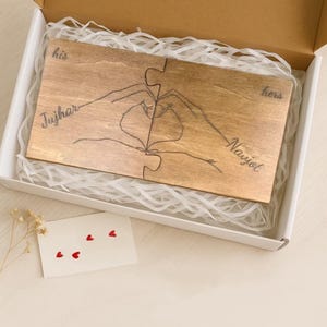 Può includere: Un puzzle in legno a due pezzi, con mani disegnate a mano che formano un cuore. Le parole "his" e "hers" sono scritte sul puzzle, insieme ai nomi "Tujhar" e "Nayjot". Il puzzle è in una scatola bianca con imbottitura di carta, ed è presente anche una carta con cuori rossi.