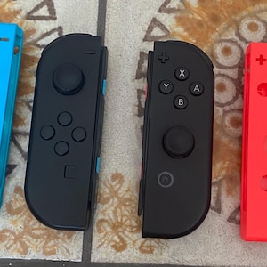 May include: Four game controllers in a row. Two are Joy-Cons, one blue and one red. The other two are black. Each controller has buttons and control sticks for gaming. The controllers are for a games console.
