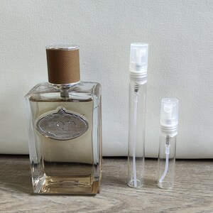 Peut inclure: Un flacon de parfum rectangulaire transparent avec un bouchon marron et une étiquette argentée portant la mention "PRADA". Deux flacons pulvérisateurs transparents sont à côté du flacon. Le parfum est de couleur ambre clair.