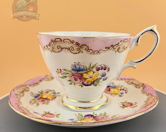 Puede incluir: Un juego de taza y platillo de té rosa y blanco con un diseño floral. La taza y el platillo tienen un borde dorado y un borde rosa.