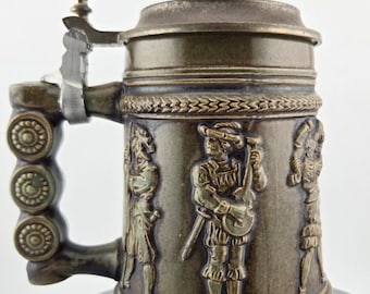 May include: A vintage beer stein with a hinged lid and handle. The stein is a muted bronze colour and features relief carvings of figures in period clothing. The handle has a decorative design.
