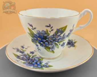Könnte beinhalten: Eine weiße Teetasse und Untertasse mit einem blauen und grünen Blumenmuster. Das Blumenmuster zeigt Vergissmeinnicht-Blumen.