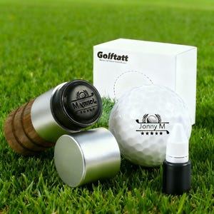 May include: A golf ball, stamp, ink bottle, and box are displayed on green grass. The golf ball has a personalized design with the name "Jonny M". The stamp has a wooden handle and a metal casing. The box is white and has the text "Golftatt".