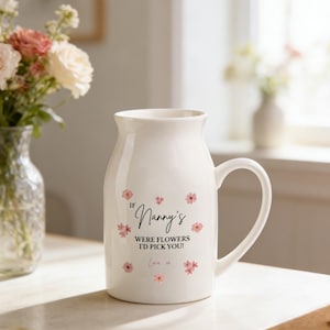 Könnte beinhalten: Eine weiße, krugförmige Keramiktasse mit Henkel, verziert mit rosa Blumenmotiven und dem Text "If Nanny's Were Flowers I'd Pick You!" in schwarzer Schrift. Die Tasse steht auf einer hellen Oberfläche, mit Blumen im Hintergrund.
