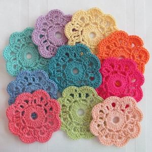 May include: A collection of ten colorful, crocheted flower-shaped coasters. The coasters are in various shades, including pink, orange, yellow, green, blue, and purple. Each coaster has a central hole and scalloped edges, creating a floral design.