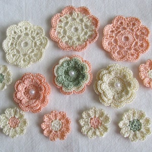 May include: A collection of handmade crochet flowers in various sizes and colors. The flowers feature shades of cream, peach, and green, with some adorned with a pearl bead in the center. The crochet work is detailed, showcasing a delicate floral design.