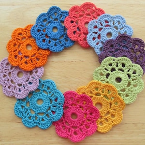 May include: A collection of colorful, crocheted flower-shaped doilies arranged in a circular pattern. The doilies are in various colors, including orange, blue, pink, purple, and green. The openwork design and vibrant hues create a cheerful, handmade aesthetic.