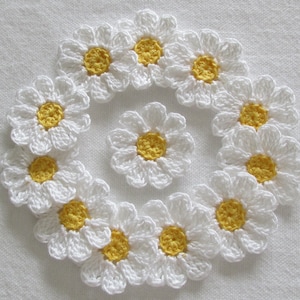 May include: A collection of handmade crochet daisies. The daisies have white petals and yellow centers. The flowers are arranged in a circular pattern, with one daisy in the center. The background is a plain white fabric.