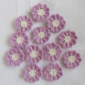 May include: Twelve crocheted lavender flowers with white centers. The flowers are arranged in a circle on a white background.