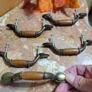 May include: Antique-style cabinet pulls with a bronze finish and wooden center. The pulls have a curved design with screw attachments. One pull is held by a hand. The pulls are on a marble-like surface.