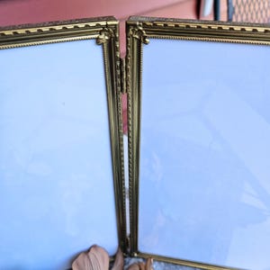 May include: A hinged, double-panel picture frame with a gold-colored metal frame. The frame has ornate detailing and a decorative floral element at the corners. The panels are empty with a white background.
