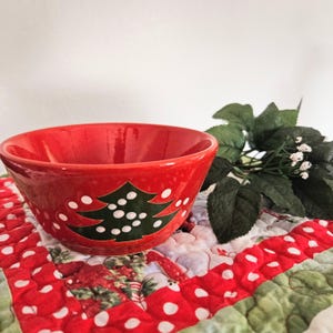 May include: A red ceramic bowl with a Christmas tree design. The bowl is round with a wide opening and a glossy finish. The Christmas tree is green with white dot ornaments. The bowl is sitting on a red and white polka dot quilted placemat.