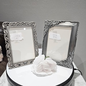 May include: Two silver-colored picture frames with ornate, detailed borders. Each frame has a white interior and a label with handwritten text. A white rose is placed in front of the frames on a white surface.