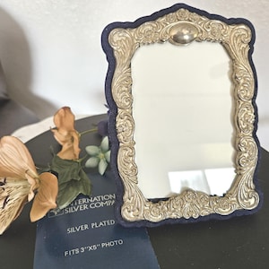 May include: Silver-plated photo frame with ornate scrollwork and a dark blue velvet border. The frame is designed to hold a 3"x5" photo. The frame has a classic, vintage aesthetic.