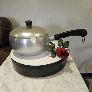 May include: A vintage aluminum saucepan with a black handle and lid. The saucepan is sitting on a white and black rotating display stand. A red rose with green leaves is placed next to the saucepan.