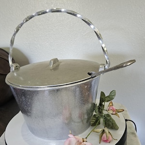 May include: A silver-colored metal serving pot with a lid and a twisted handle. A matching spoon rests on the rim. The pot is sitting on a white surface with pink flowers.