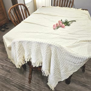 May include: Cream-colored textured tablecloth with fringe trim, draped over a wooden table. A small bouquet of pink roses rests on the tablecloth. The table is surrounded by wooden chairs.
