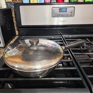 May include: A stainless steel frying pan with a lid, sitting on a black gas stove. The pan has a black handle and a round, dark knob on the lid. The stove has a digital display showing the time.