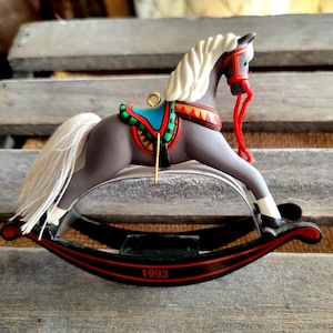 May include: A gray rocking horse ornament with a white mane and tail, featuring a teal and red saddle, a red bridle with matching reins, and a black rocker base with red stripes. The year 1993 is printed in red on the front of the rocker base.