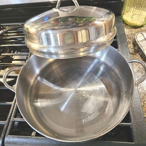 May include: A stainless steel pot with a matching lid, resting on a black stovetop. The pot has two curved handles and a shiny, reflective surface. The lid is slightly ajar, revealing the interior of the pot. Kitchenware.