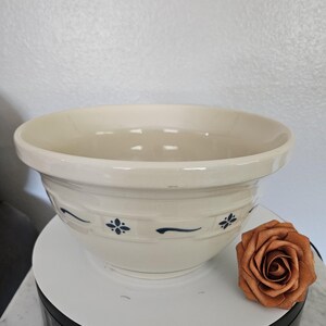 May include: A cream-colored ceramic mixing bowl with a wide rim and a decorative embossed band featuring dark blue floral and curved line patterns. The bowl has a smooth, glossy finish and a deep, rounded interior, presented with a copper-colored rose.