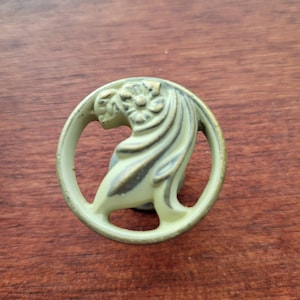 May include: A vintage-style, circular cabinet pull with a floral and flowing design. The metal hardware has a distressed, antique finish with shades of gold and green. The decorative element is set within a circular frame.
