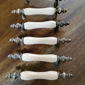 May include: Six drawer pulls arranged in a vertical row. Each pull features a smooth, cream-colored ceramic center handle flanked by ornate, antique-style brass metal ends with a detailed, multi-tiered finial design on both sides.
