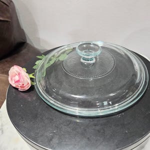 May include: A clear glass lid with a circular design and a small handle. The lid has a series of concentric ridges radiating from the center. A pink rose and green leaves are on the left side of the lid. The lid is on a black surface.