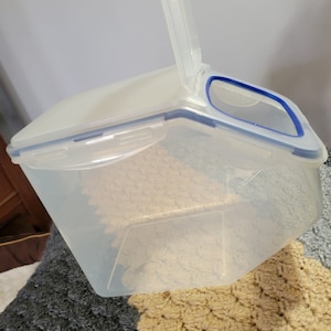 May include: A clear plastic food storage container with a blue and white lid and handle. The container is filled with a beige substance. The container is sitting on a gray and beige crocheted blanket.