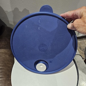 May include: A round, navy blue Tupperware lid with a small handle and a white circular valve. The lid has a textured surface and the Tupperware logo is visible. The lid is being held by a person's hand.