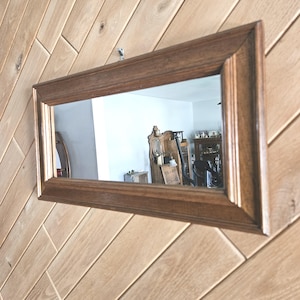 May include: Rectangular wooden framed mirror. The mirror has a dark brown frame and is hanging on a light brown wooden wall. The mirror reflects a room with furniture.