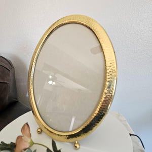May include: Oval gold-colored picture frame with a hammered texture. The frame has a glass insert and is supported by three small, gold-colored ball feet. The frame is empty, ready to display a photograph or artwork.