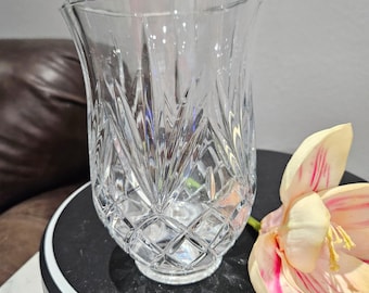 May include: A clear, cut-glass vase with a flared top and diamond and vertical line patterns. The vase sits on a black surface, with a partially visible flower with pink and yellow petals to the right. The vase is approximately 7 inches tall.