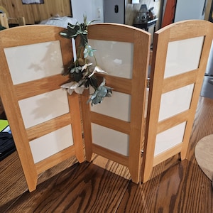 May include: A three-panel folding photo frame made of light-toned wood with white rectangular inserts in each section. The frame is hinged to stand upright and is decorated with a small cluster of artificial green leaves and white flowers.