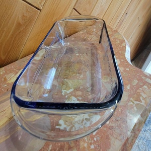 May include: A clear, rectangular glass baking dish with rounded corners. The dish has a blue tint and decorative etched patterns along the sides. The dish is sitting on a marble surface.