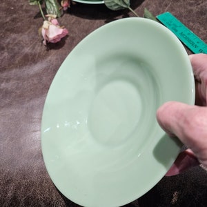 May include: A light green, oval-shaped saucer with a slightly raised rim. The saucer is held by a hand, with a brown surface in the background. A dried rose and a ruler are also visible.